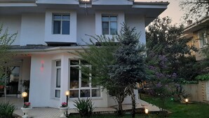 Une maison à deux étages avec un balcon, une antenne parabolique et un jardin où poussent différentes plantes et fleurs.