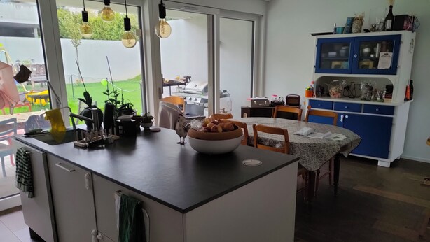 A modern kitchen with a central island, a dining area with a table, and a blue cabinet with glass doors. - Room in our family home, close to the station and services (Courtepin)