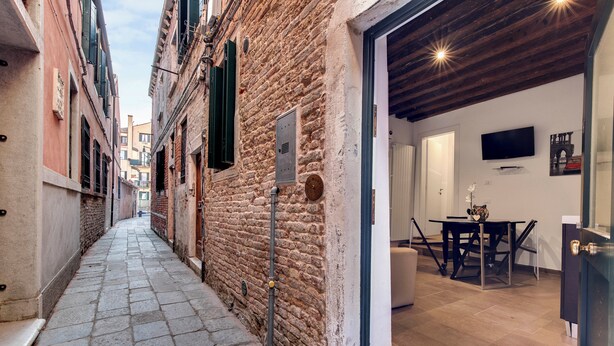 A narrow alley with stone pavement, a brick wall, and a wooden ceiling. There is a door leading to a room with a television and a table. - I Quattro Mori by Wonderful Italy (Venice)