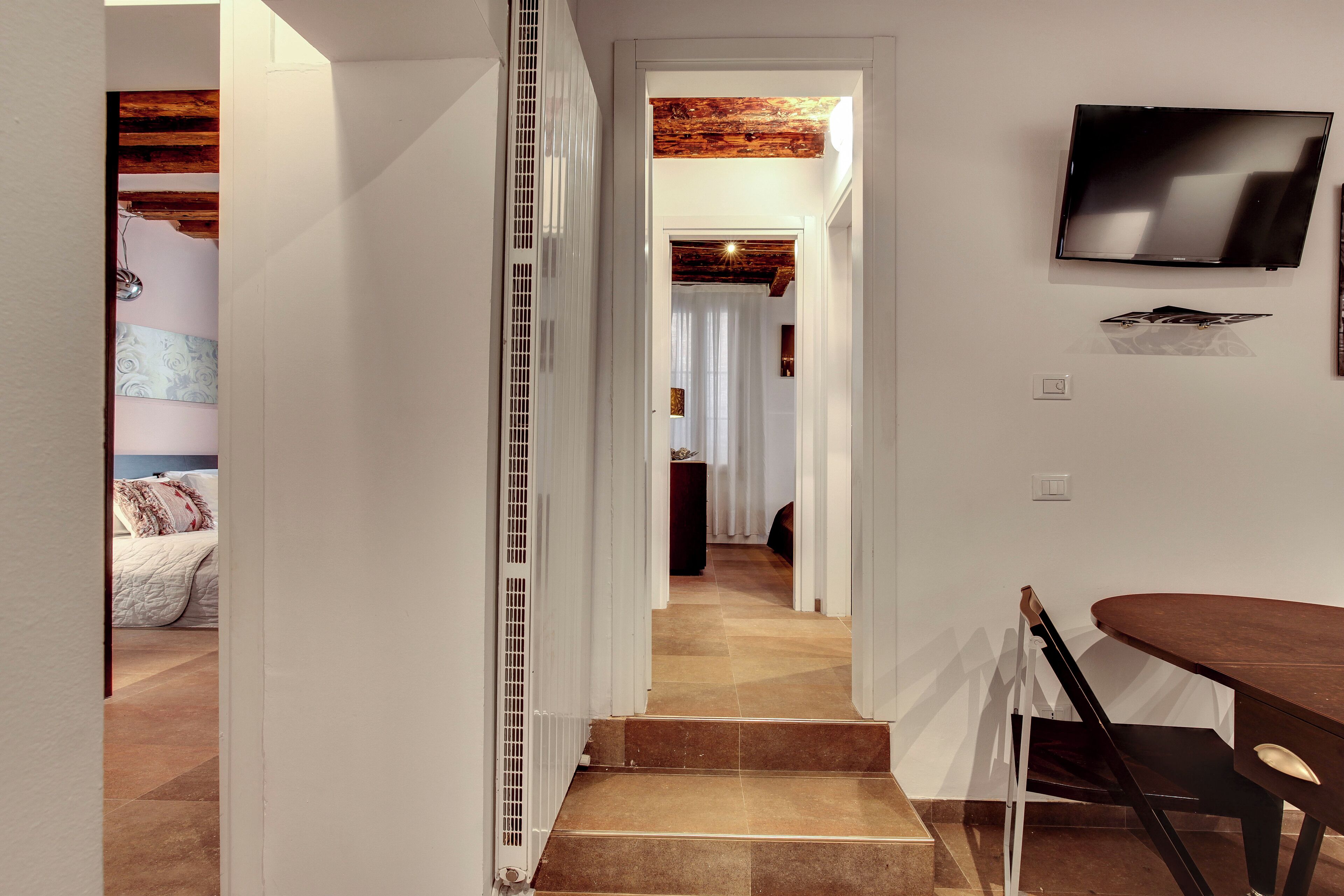 A hallway with a staircase leading to a bedroom, a dining area with a table and chairs, and a mounted television on the wall.