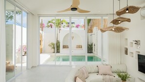 A modern living room with a large sofa, a coffee table, and a TV mounted on the wall. There is a large window with a view of a pool and outdoor plants. - New 1BR Private Pool Villa in Bingin, Uluwatu (Bali)
