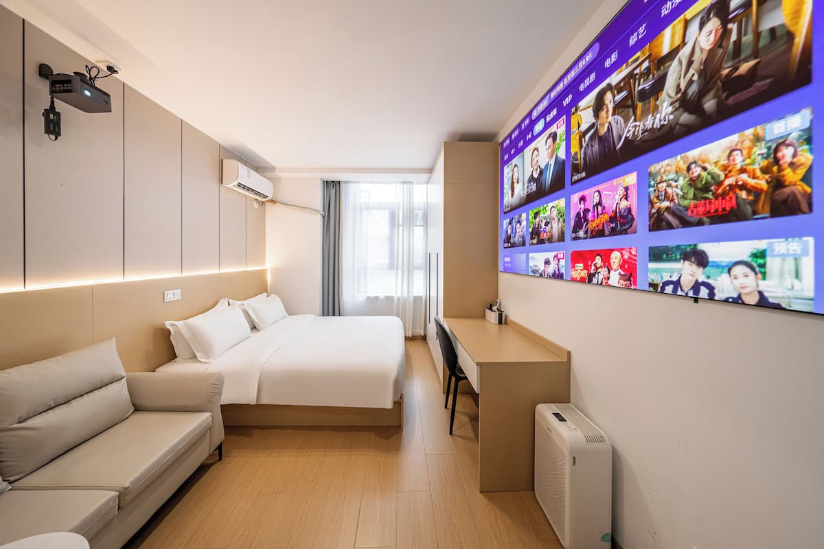 A modern hotel room with a bed, a desk, a sofa, and a wall-mounted TV displaying various shows.