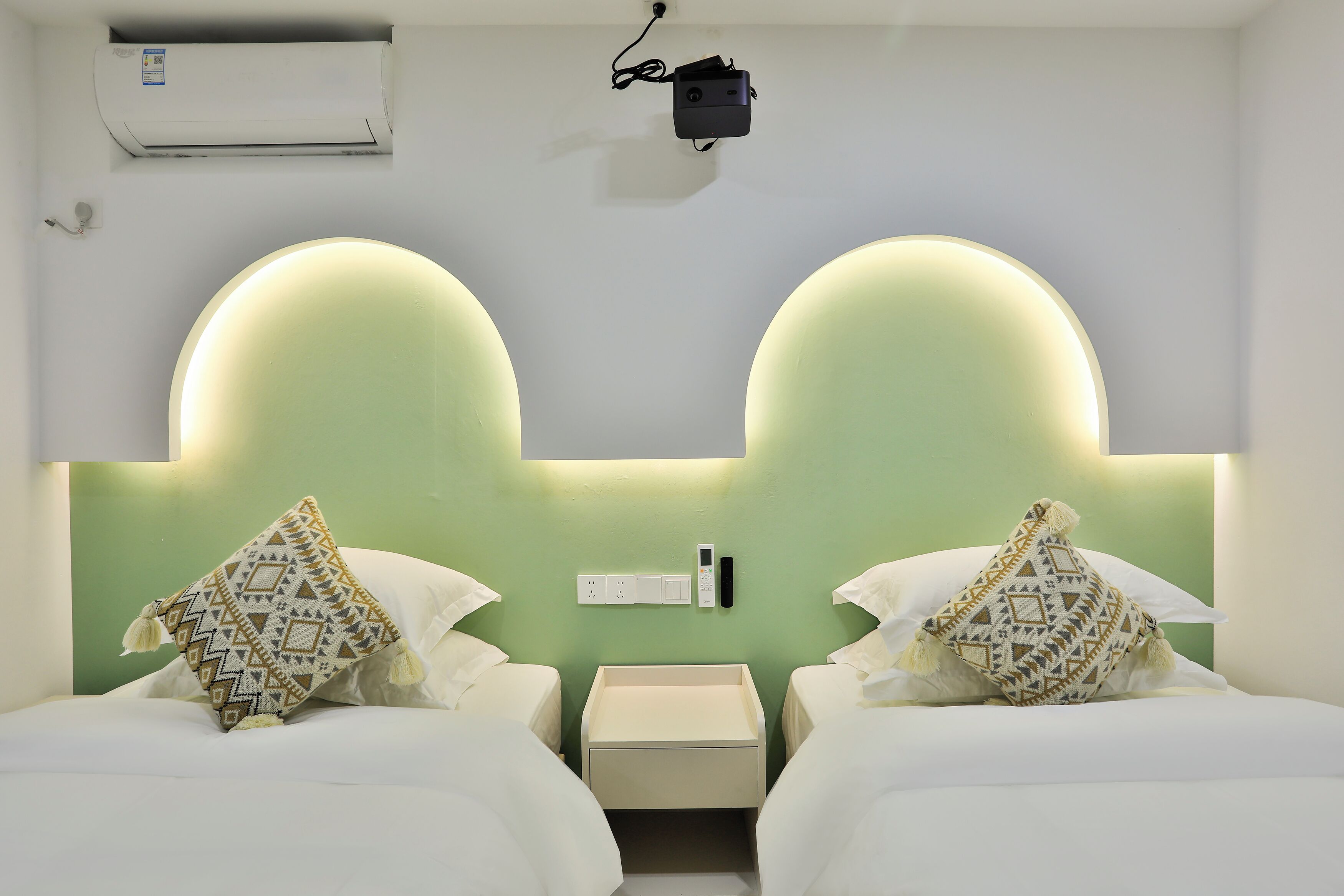A hotel room with two beds, a bedside table, a nightstand, a wall-mounted air conditioner, and a wall-mounted projector.