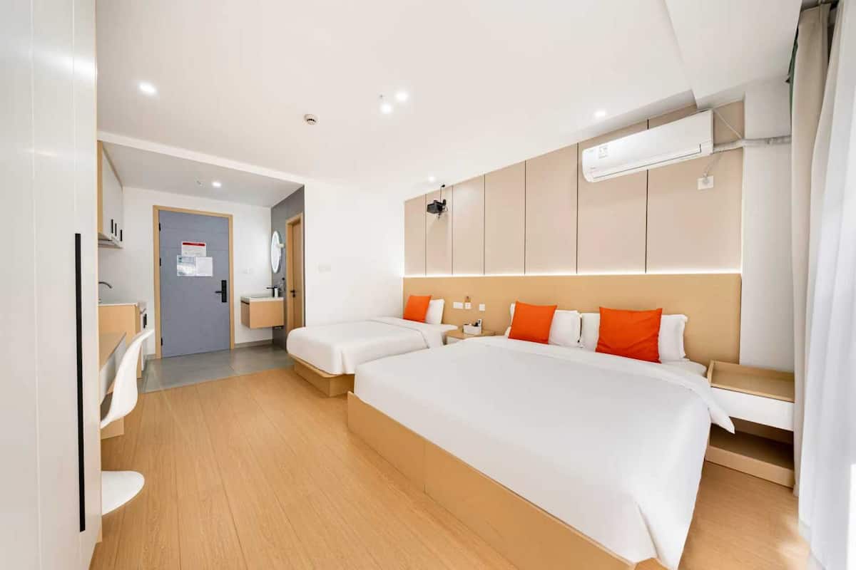 A hotel room with a large bed, a bedside table, a small desk, a chair, a mirror, a wardrobe, and an air conditioning unit.