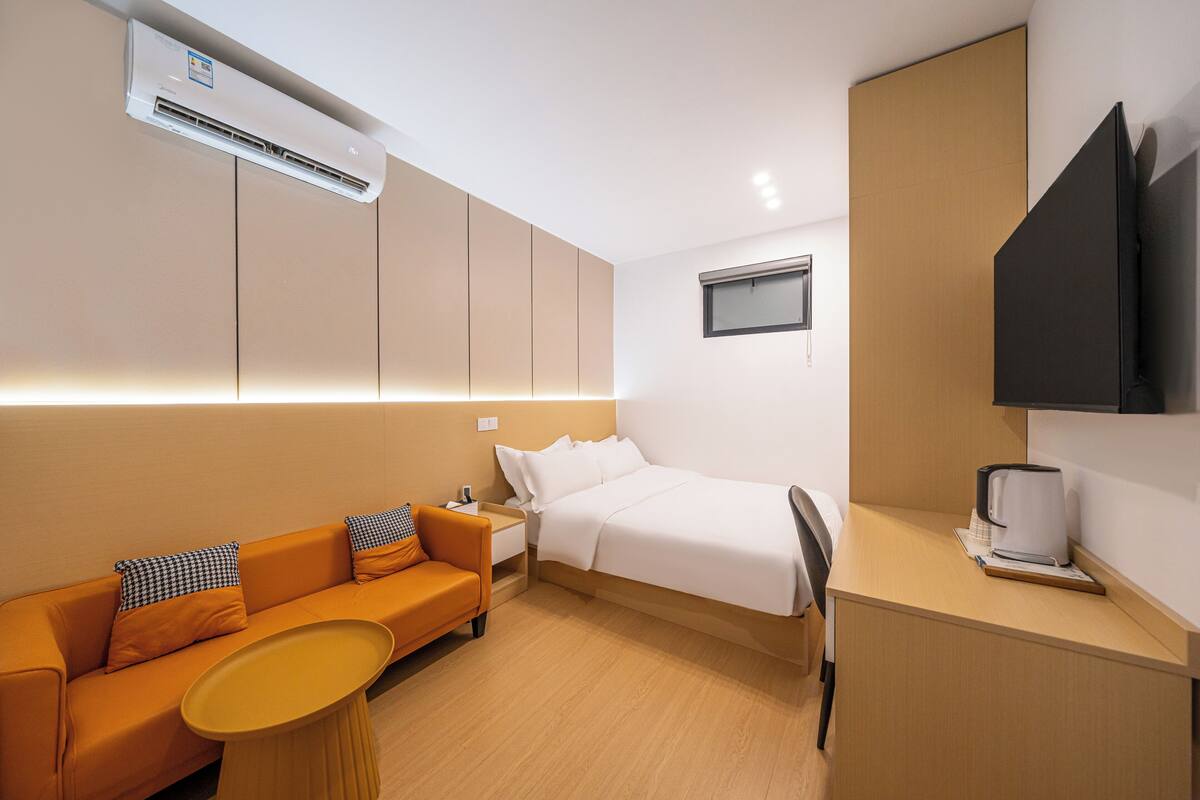 A modern hotel room with a bed, a sofa, a small table, a TV, and an air conditioning unit.