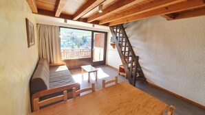 Ein gemütliches Zimmer mit Holzbalkendecke, einer Couch, einem Esstisch und Blick auf eine Berglandschaft durch ein großes Fenster.