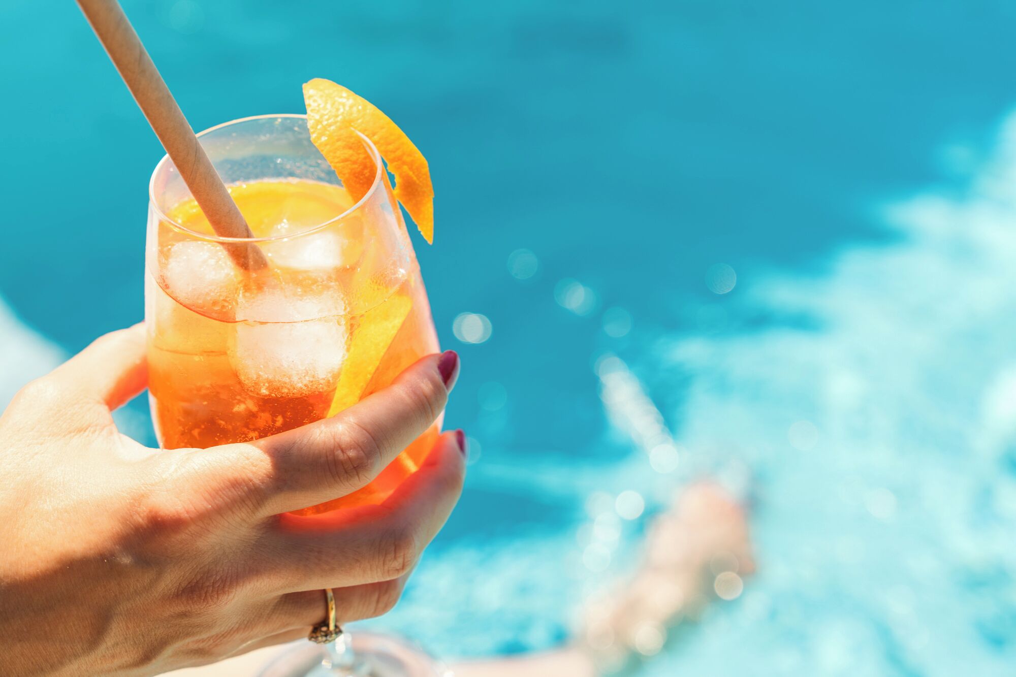 A hand holding a glass with a straw and an orange slice, with a pool in the background.