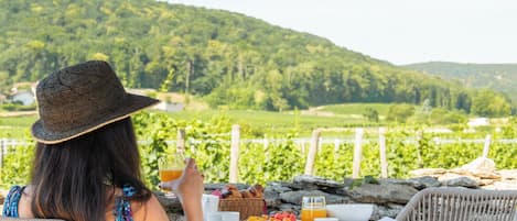 A person seated at an outdoor table with a view of a vineyard and hills in the background.