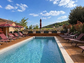 A rectangular swimming pool with lounge chairs and umbrellas on a wooden deck, surrounded by greenery and houses in the background. - Hotel Les Deux Chevres Gevrey-Chambertin (Gevrey-Chambertin)