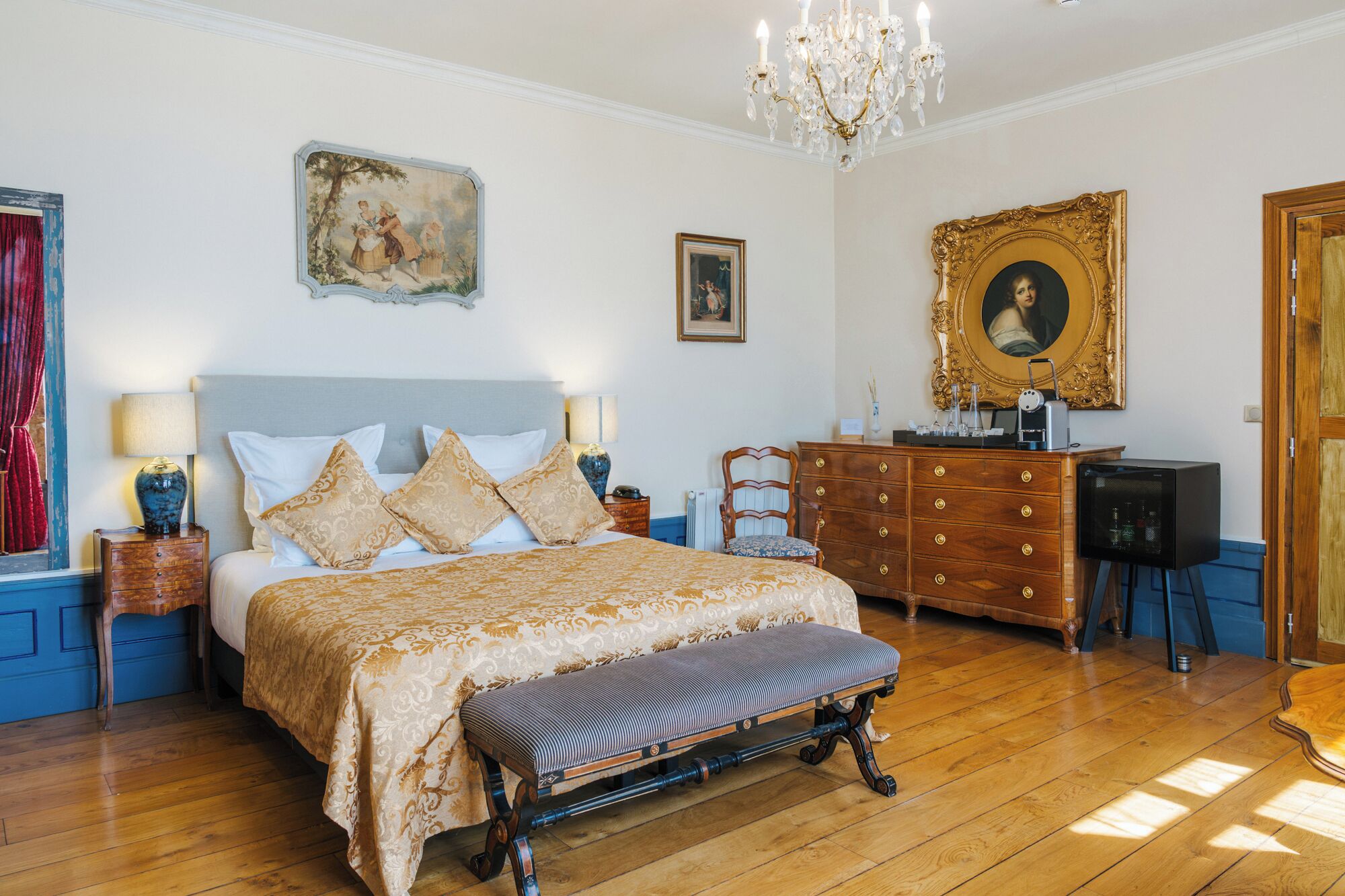 Un dormitorio con una cama grande, un aparador, un banco, una araña y obras de arte en las paredes.