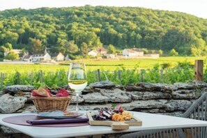 Mesa servida con una copa de vino blanco, una cesta de pan y un plato de comida, con vistas a un viñedo y casas.