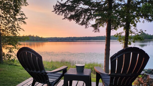 A wooden deck with two Adirondack chairs and a small table, overlooking a calm lake with trees in the background. - Private lake cabin. WiFi, Beach, Pontoon Rental. Booking for summer of 27! (Nevis)