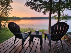 Une terrasse en bois avec deux fauteuils Adirondack et une petite table, donnant sur un lac paisible, avec des arbres en arrière-plan.