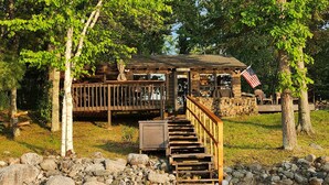 A rustic wooden cabin with a porch and a small deck, surrounded by trees and a sandy area with rocks. - Private lake cabin. WiFi, Beach, Pontoon Rental. Booking for summer of 27! (Nevis)