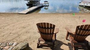 A beach with two wooden Adirondack chairs, a dock with a covered boat, and a calm lake with trees in the background. - Private lake cabin. WiFi, Beach, Pontoon Rental. Booking for summer of 27! (Nevis)