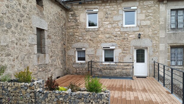 A stone building with multiple windows and a door, a wooden deck, and a stone wall with plants. - Quiet country house  (Le Buisson)