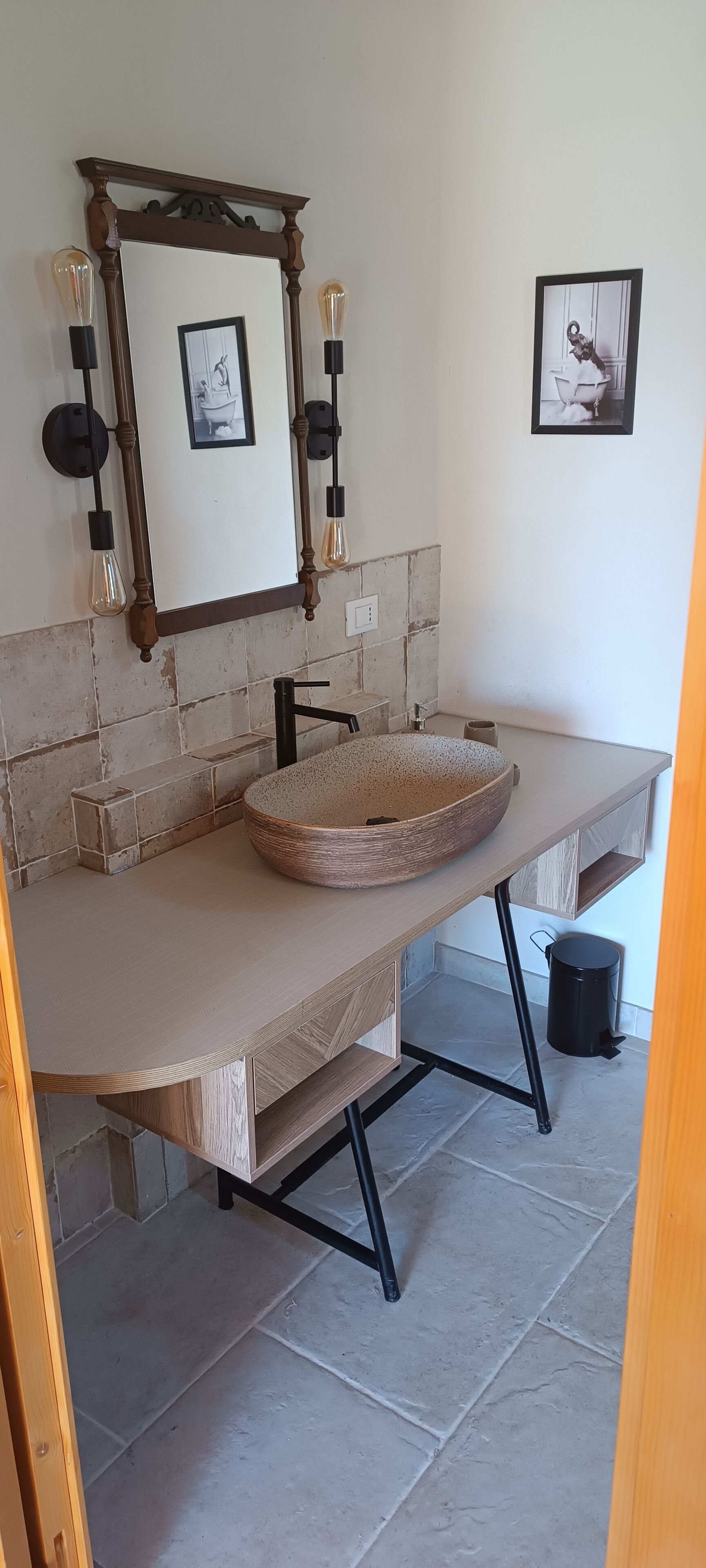 A bathroom with a round, copper-colored sink, a wooden vanity, and a mirror with a decorative frame. There are two wall-mounted light fixtures and two framed pictures on the wall.