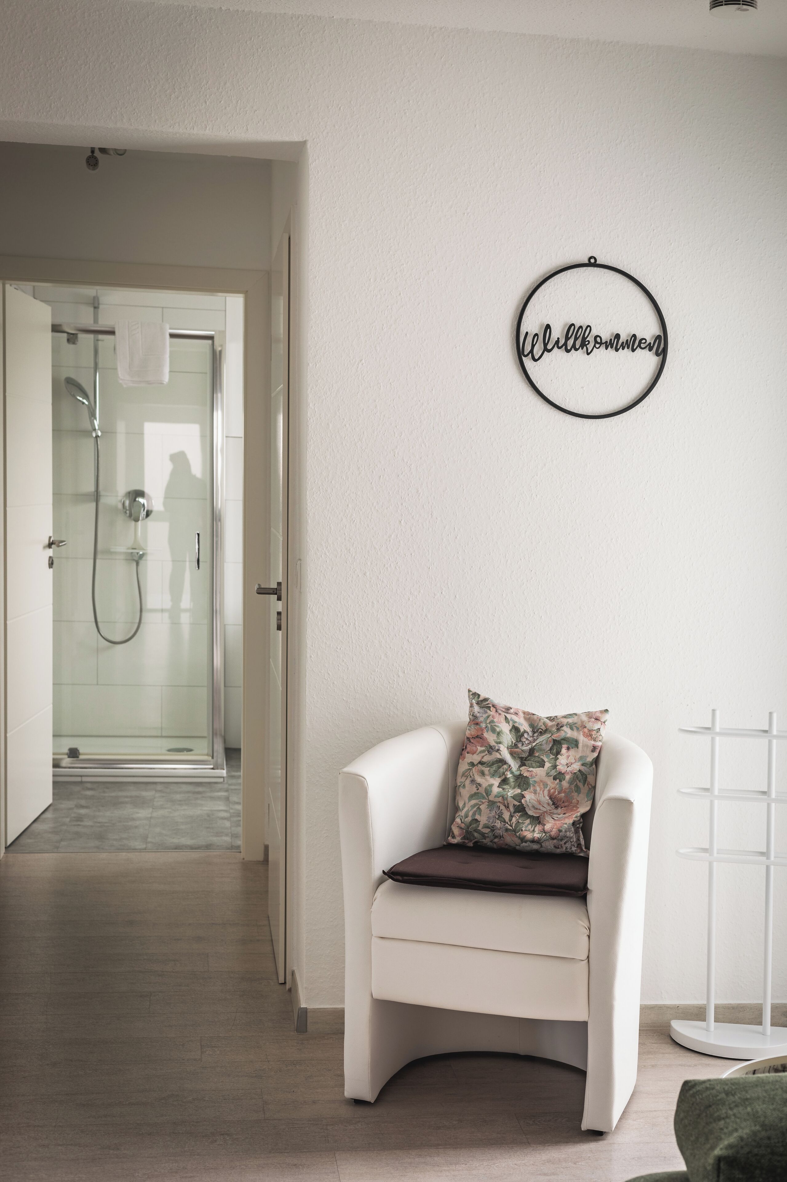 A modern bathroom with a glass shower enclosure, a white chair with a floral pillow, and a wall-mounted sign that reads "Welcome".