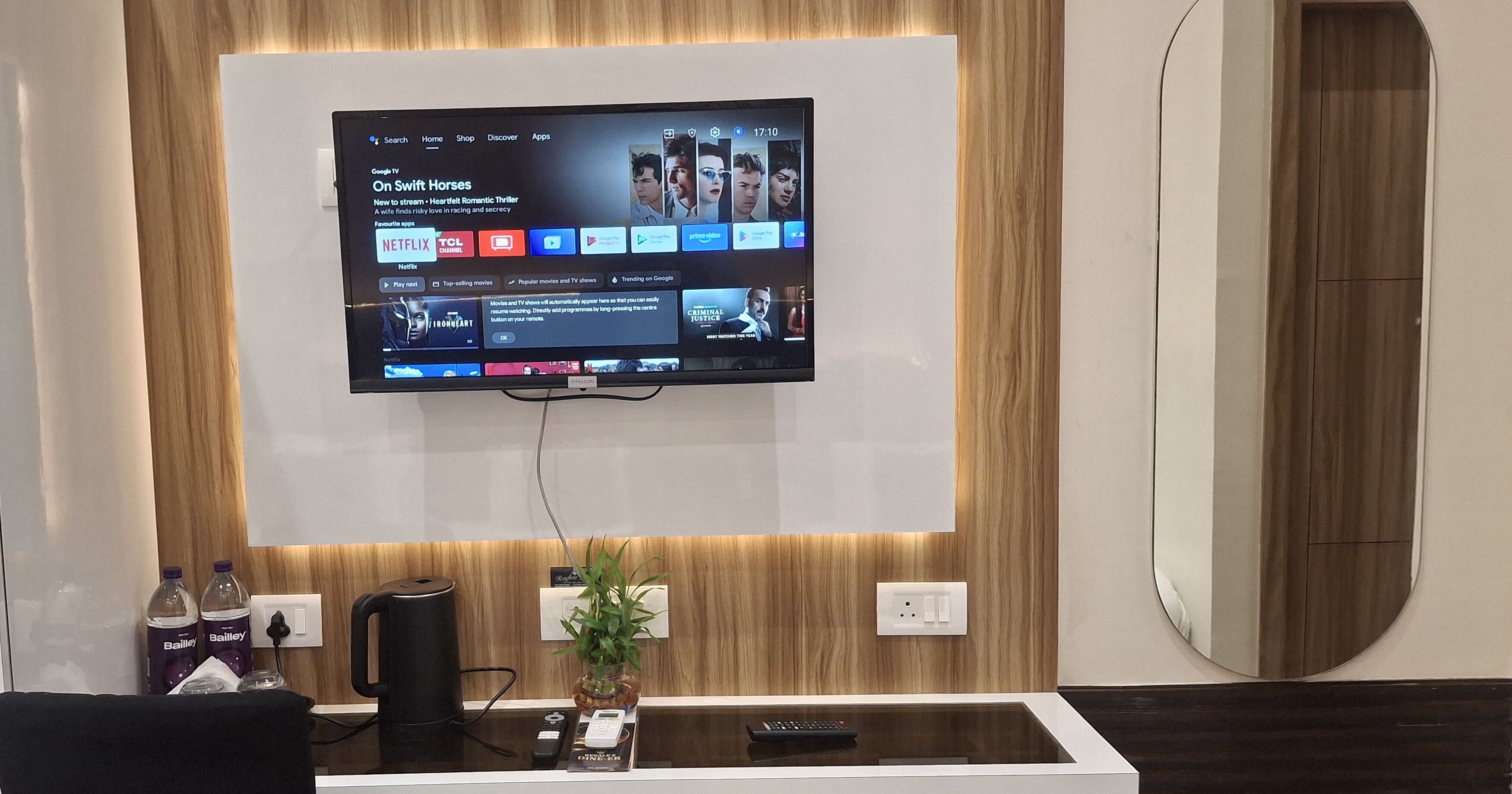 A modern hotel reception area with a mounted TV displaying streaming services, a reception desk with a plant, and a mirror reflecting the room.