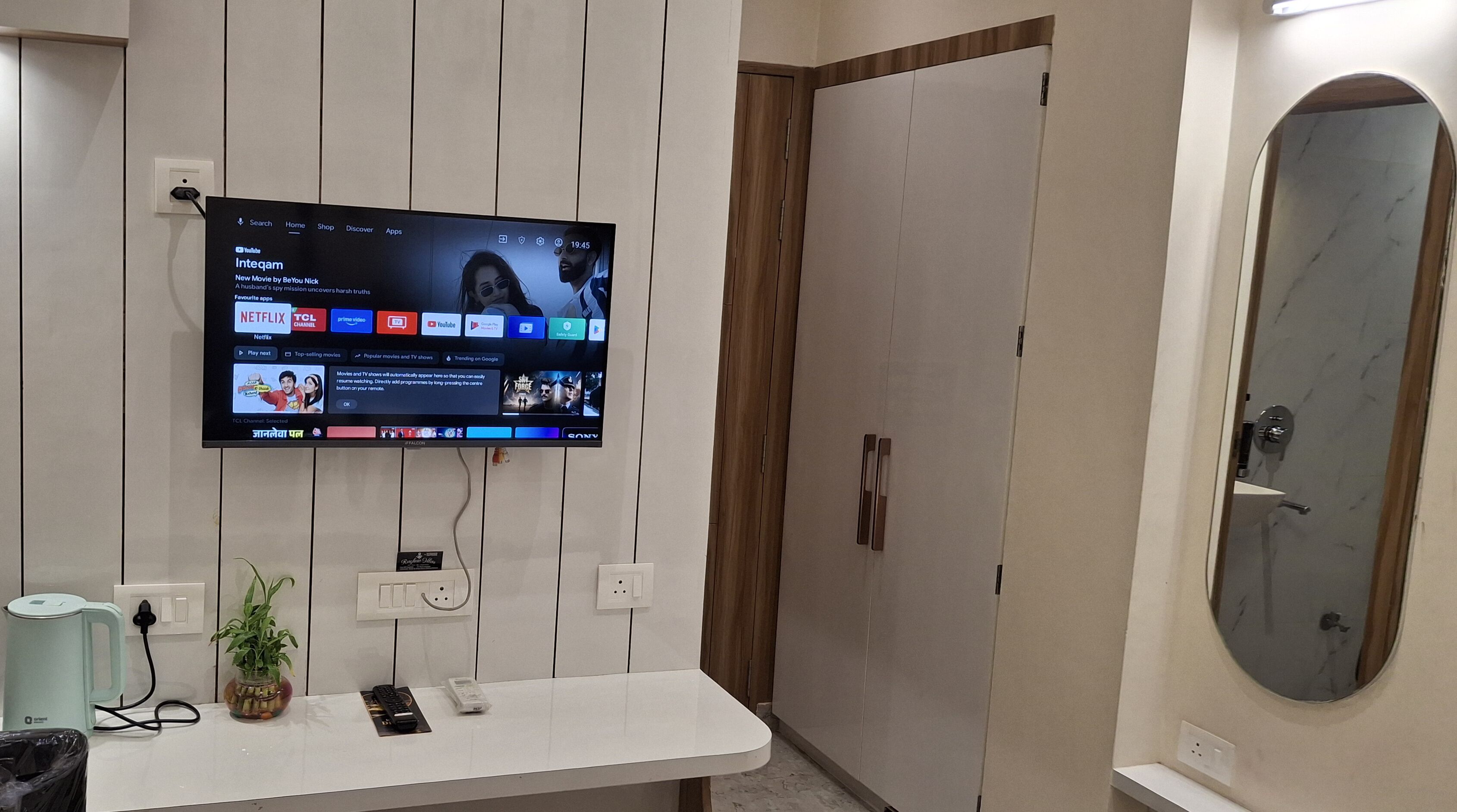 A modern bathroom with a mounted TV displaying various streaming apps, a white sink, a potted plant, and a mirror.