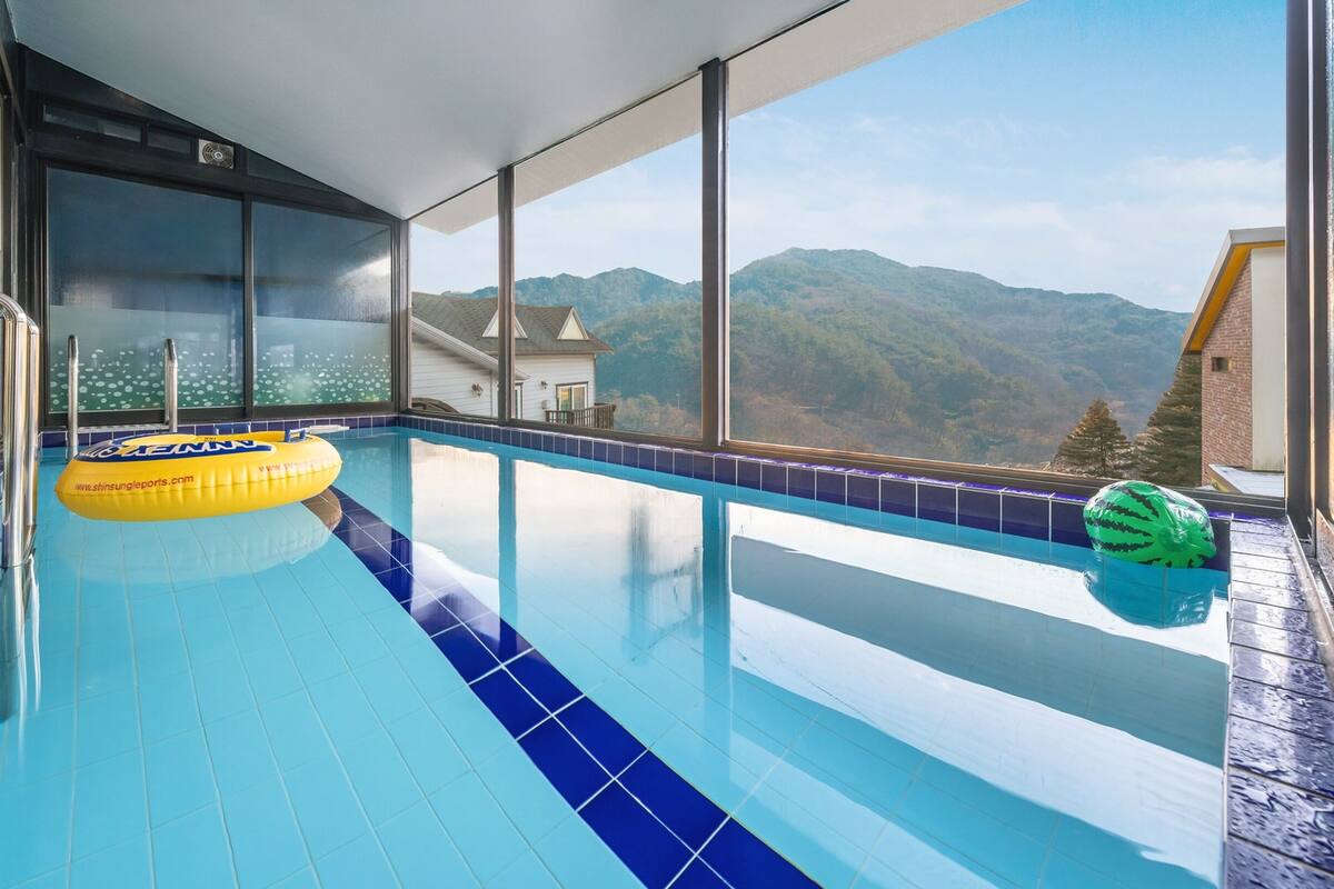 Une piscine intérieure moderne, avec des carreaux bleus transparents, un flotteur jaune et une vue sur les montagnes à travers de grandes fenêtres.