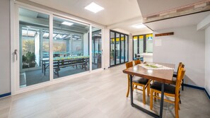 Una habitación amplia con una mesa grande y sillas, un cerramiento de vidrio corredero que da a un espacio exterior, y una pequeña cocina americana.