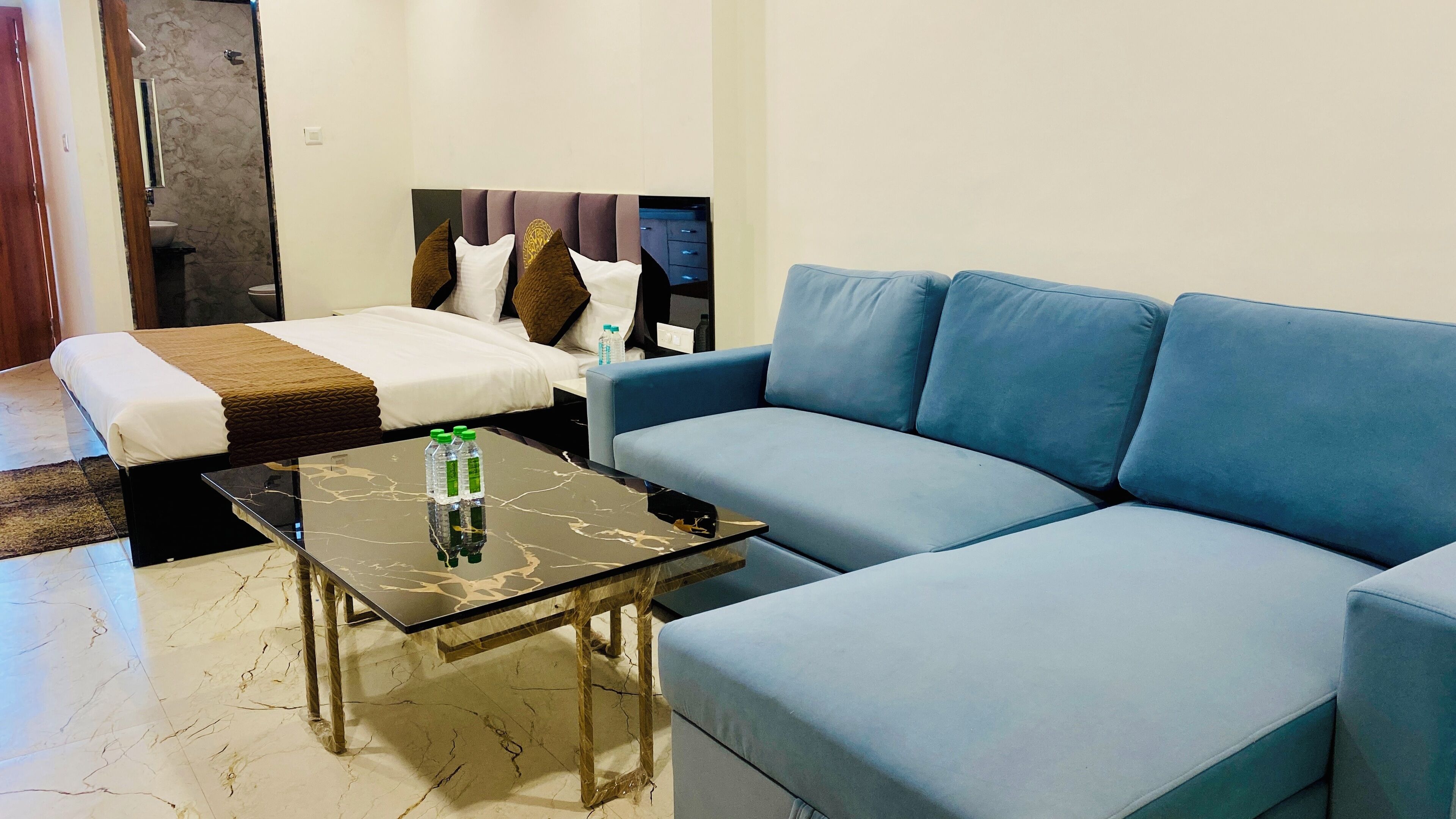 Une chambre d’hôtel avec un lit, un canapé bleu et une table basse en verre sur laquelle sont posées des bouteilles d’eau.