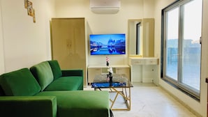 A modern living room with a green sofa, a TV mounted on the wall, a desk with a computer, and a large window offering a view of the city. - Stay 10 Parallel Apartment Hotel (Indore)