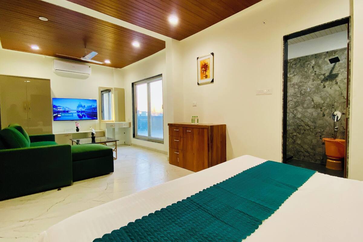 A modern hotel room with a large bed, a green sofa, a wooden cabinet, a TV, and a bathroom with a stone wall.