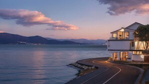 Una casa moderna de dos pisos con un camino curvo que conduce a una playa privada, con vista a un lago tranquilo y montañas al fondo.