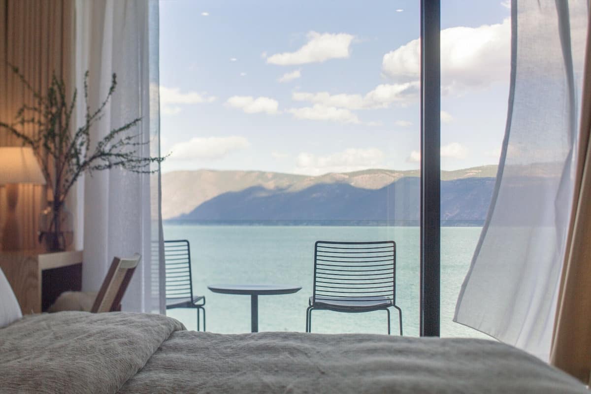 A bedroom with a large window overlooking a lake and mountains, two chairs, a bed, and a lamp.