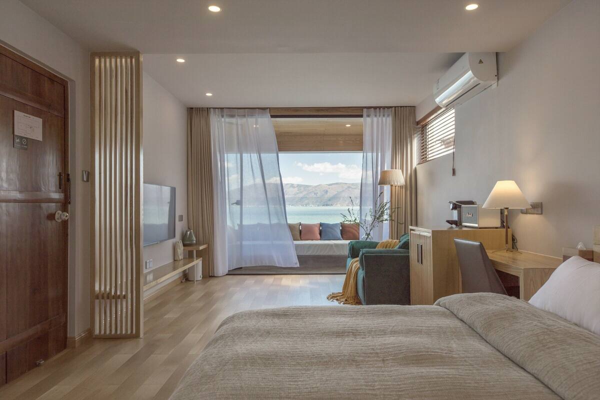 A hotel room with a bed, a desk, a chair, a TV, and a view of the sea.
