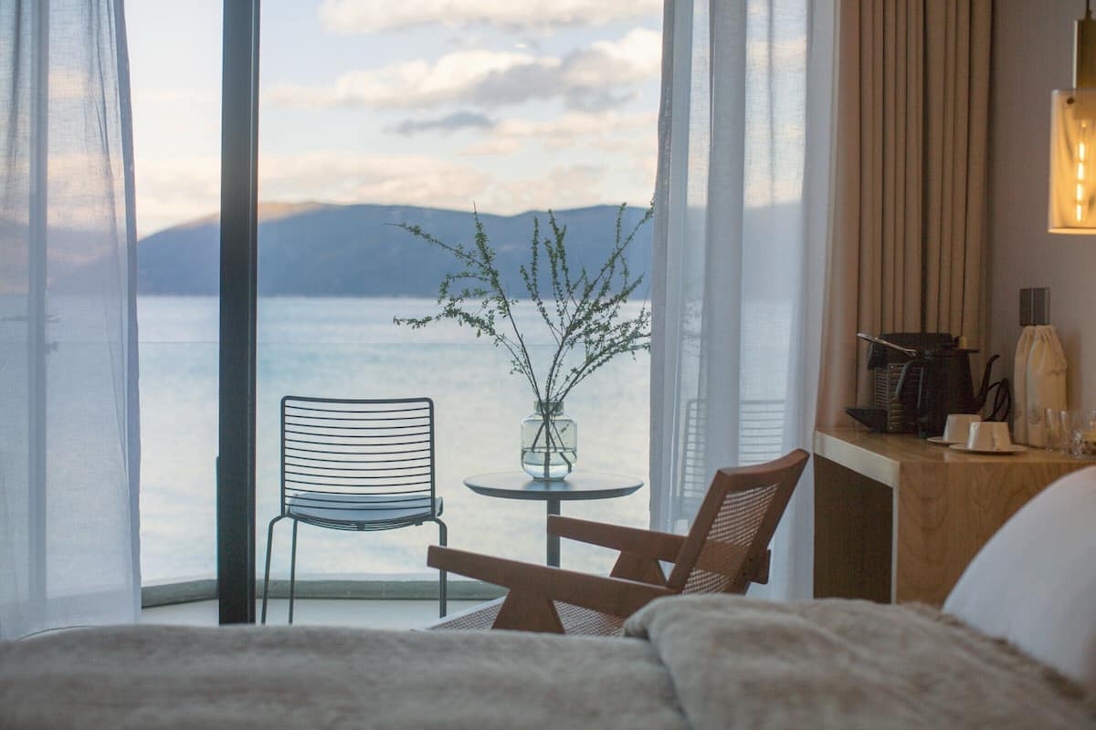 A hotel room with a bed, a desk, a chair, a small table with a vase, and a view of the sea and mountains.