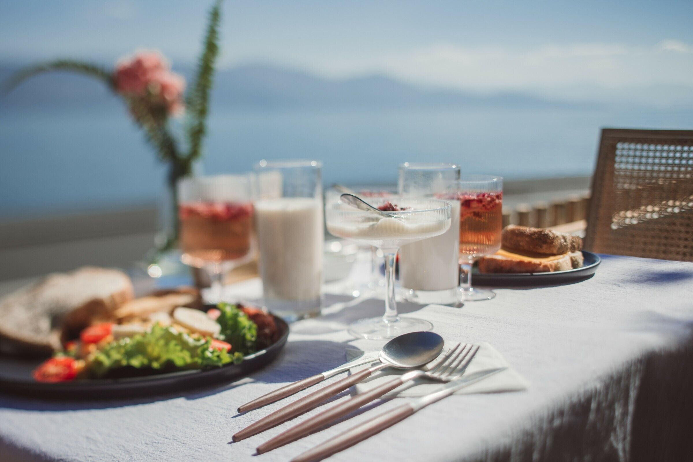 A table set with a meal, including bread, salad, and drinks, with a view of the sea and mountains in the background.