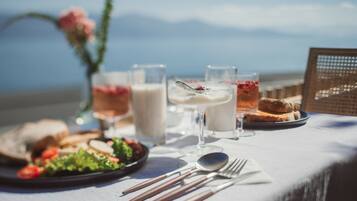 Una mesa dispuesta con comida, incluyendo pan, ensalada y bebidas, con vista al mar y montañas al fondo.