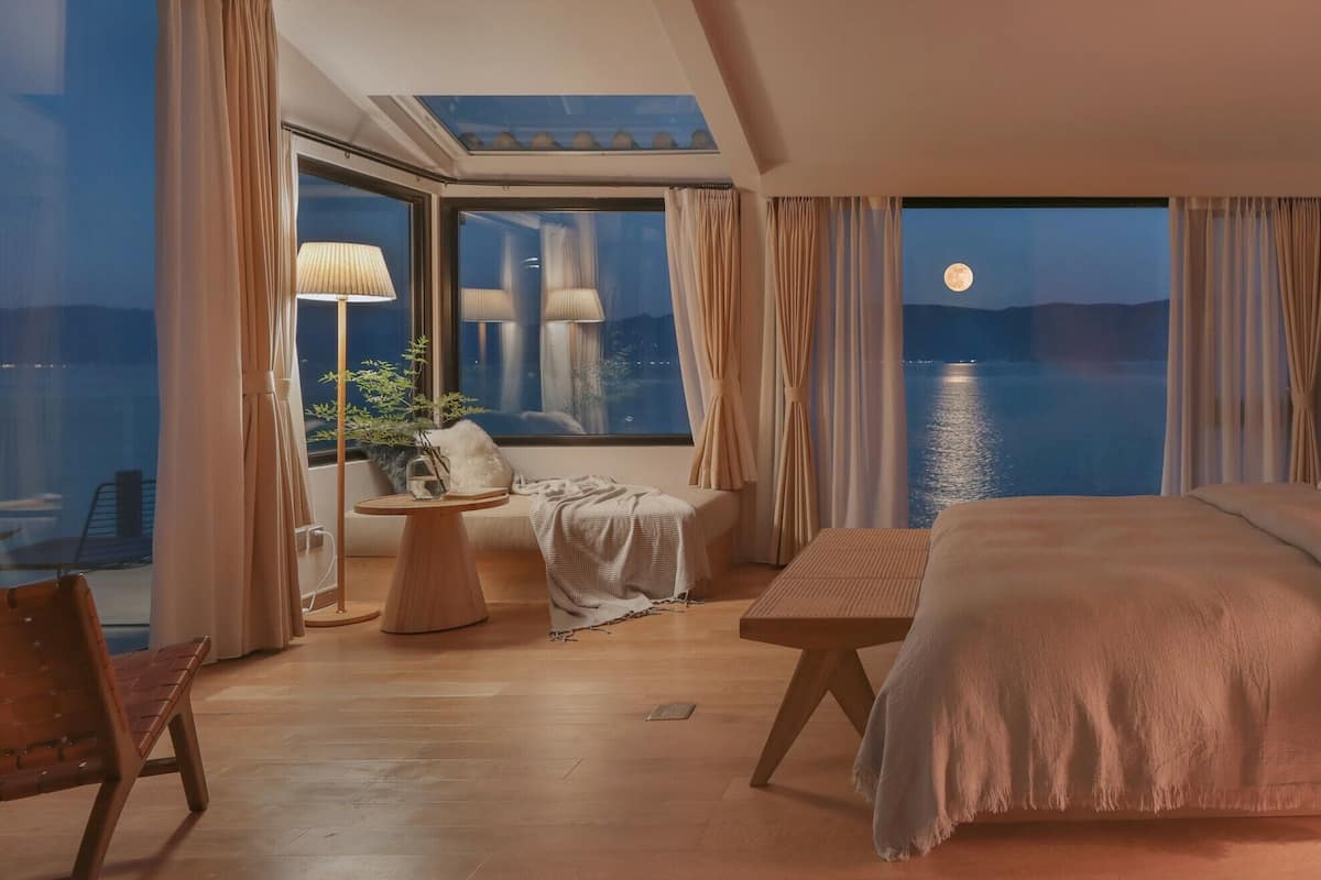 A bedroom with a large bed, a bedside table, a chair, and a window overlooking a body of water.