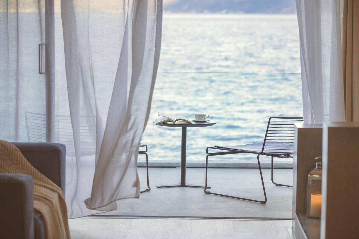 A room with a view of the sea, a table with a book and a cup, and a chair.
