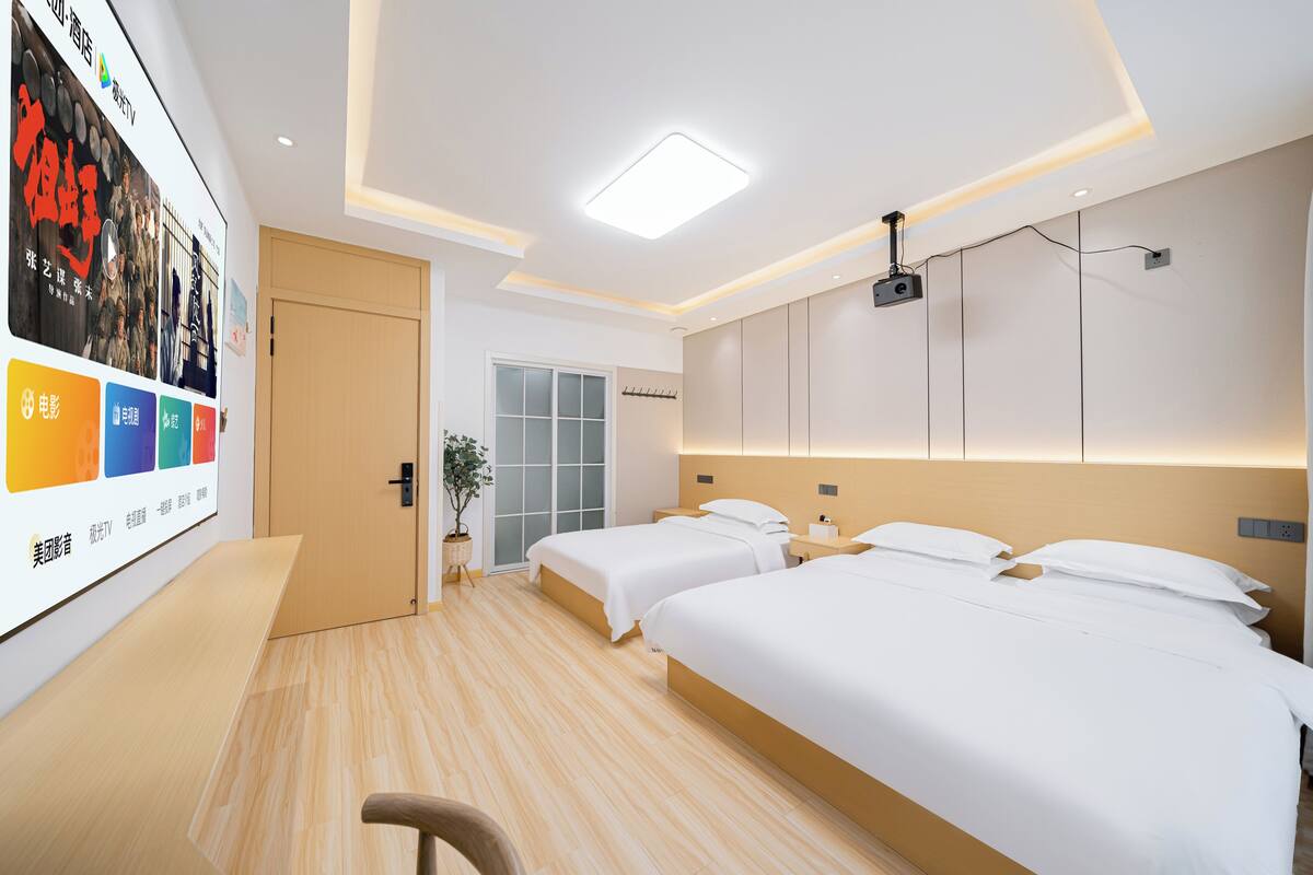 Quarto de hotel com duas camas, uma escrivaninha, um projetor e um cartaz grande na parede.