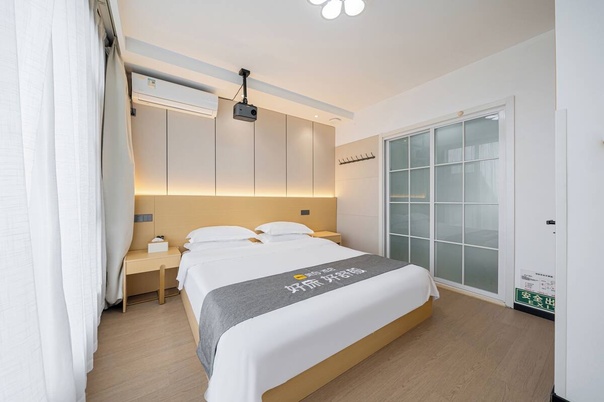Quarto de hotel moderno com uma cama bem arrumada, mesa de cabeceira, ar condicionado fixado na parede e um guarda-roupa com porta de vidro deslizante.