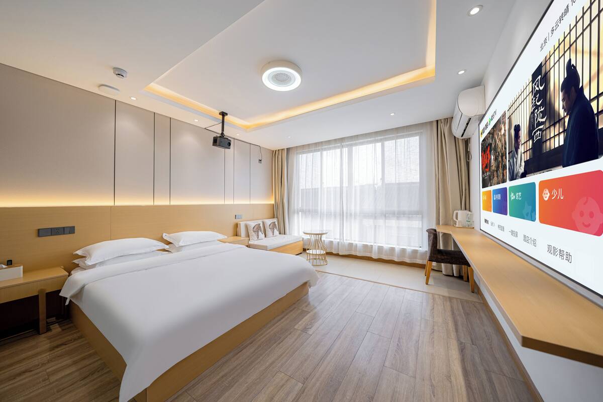 Quarto de hotel moderno com uma cama grande, mesas de cabeceira, uma escrivaninha e um janelão com cortinas.