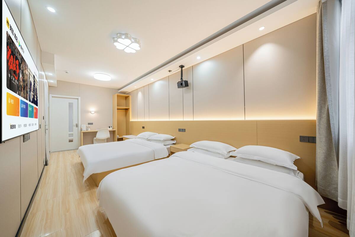 Quarto de hotel com duas camas, projetor no teto e piso de madeira.