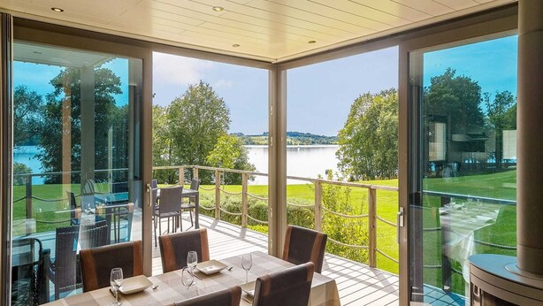 A dining area with a table set for two, overlooking a lake and grassy landscape. - Lakeside Stay with Sauna (Boussu-lez-Walcourt)