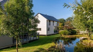 Une maison moderne dotée d’une terrasse, entourée d’une végétation luxuriante et bordée d’un étang.