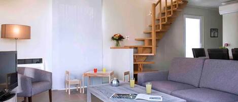 A modern living room with a grey sofa, a coffee table, a TV, a wooden staircase, and a small dining area.