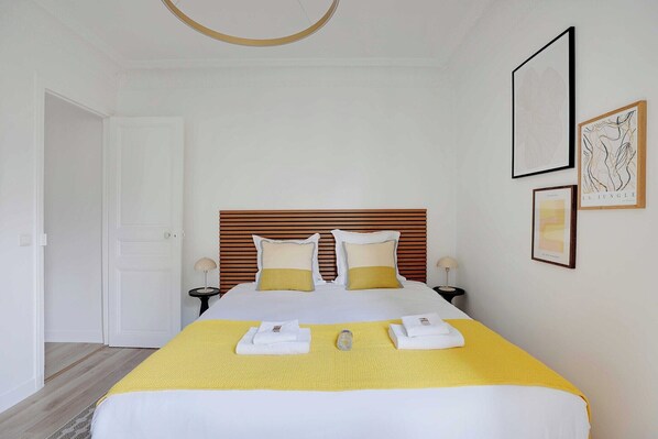 A neatly made bed with a yellow bedspread, two pillows, and two folded towels. There are framed pictures on the wall, a round ceiling light, and a wooden headboard.