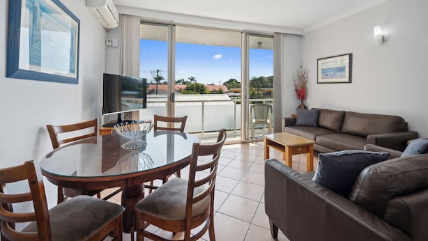 A modern living room with a round dining table, comfortable seating, a flat-screen TV, and a balcony with a view.