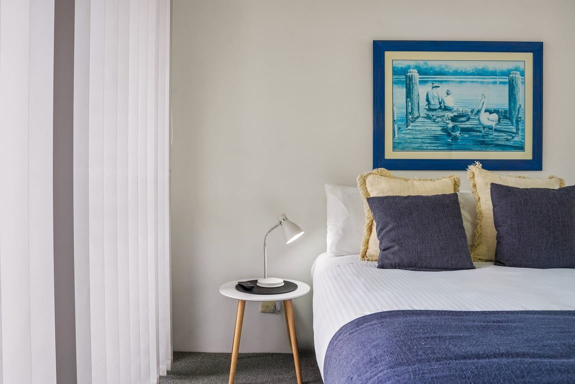 A bedroom with a bed, a nightstand with a lamp, a framed picture on the wall, and a window with blinds.