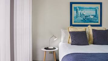 A bedroom with a bed, a nightstand with a lamp, a framed picture on the wall, and a window with blinds.