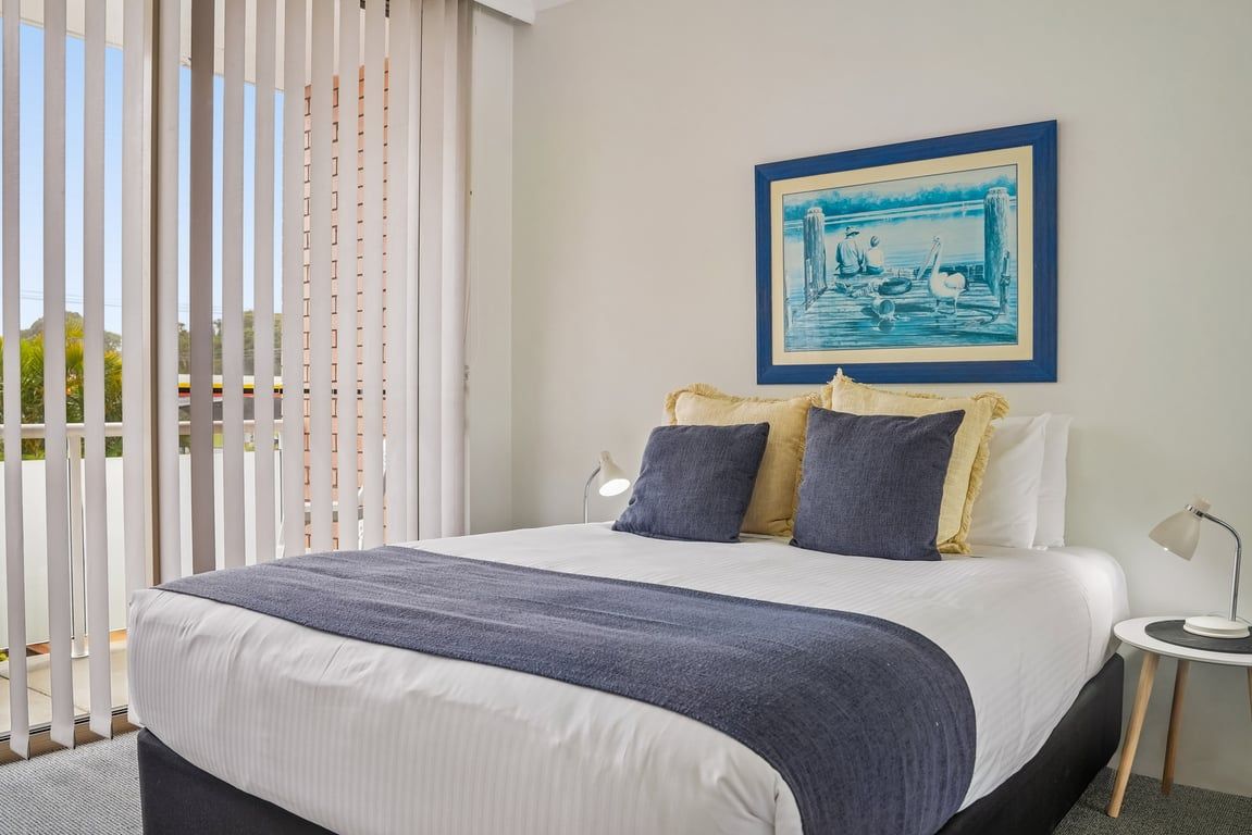 A hotel room with a bed, a framed picture on the wall, and a window with blinds.