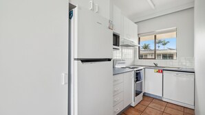 Een kleine keuken met witte kasten, een inbouwoven, een vaatwasser en een raam met uitzicht op palmbomen.